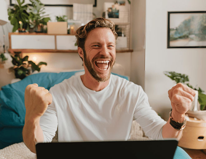 A white man cheers in his cozy room, laptop glowing with victory, fist raised in triumph, and a wide smile.