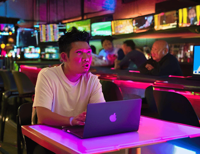 An Indonesian man, engrossed in his laptop, sits at a high table in a sports bar. Bright neon lights and multiple TV screens illuminate his face.